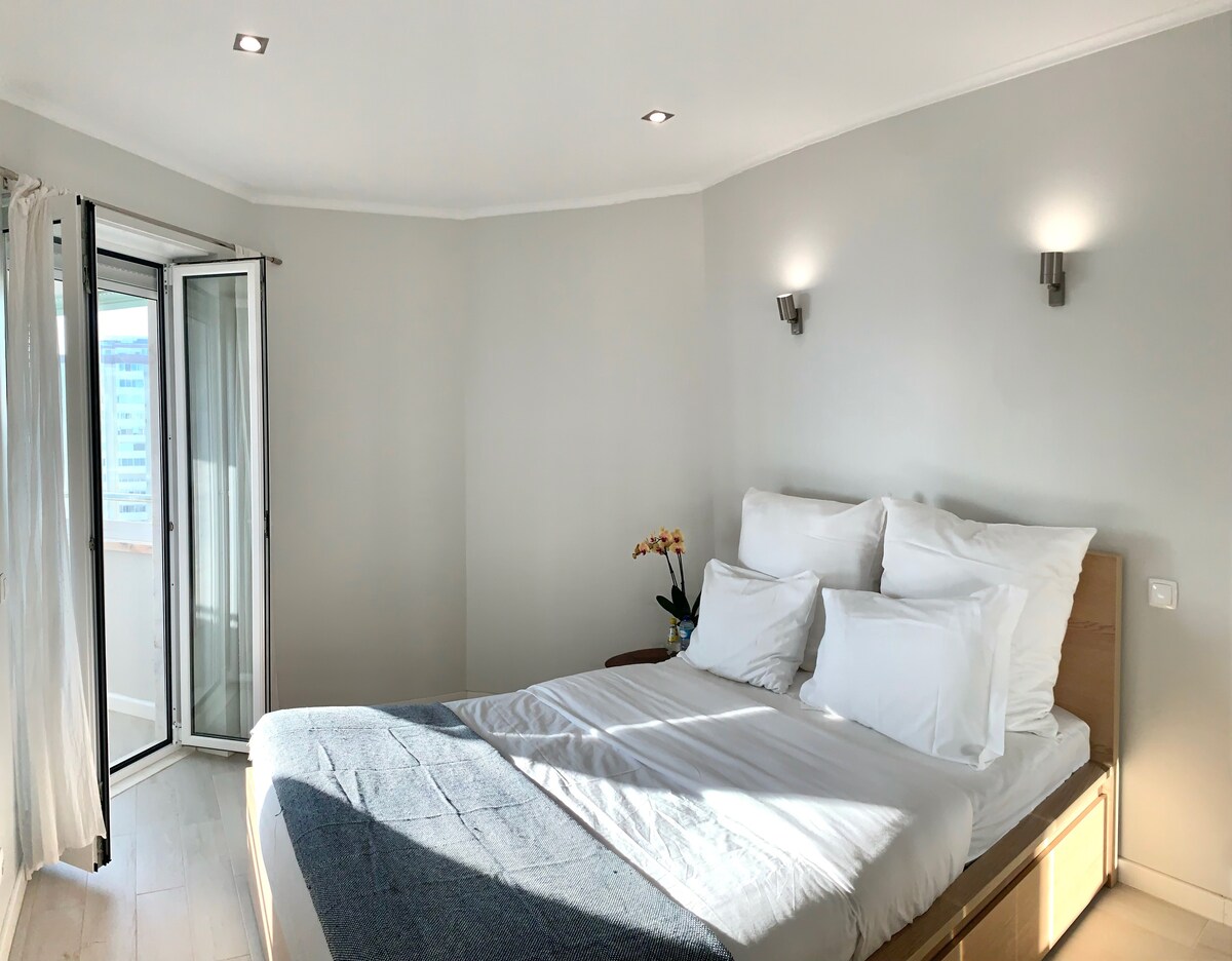A bright bedroom is presented with a spacious bed dressed in white linens, complemented by soft pillows. Natural light enters through a glass door that opens to a balcony, enhancing the calm atmosphere. Light-colored walls and two wall sconces contribute to the room's contemporary design.