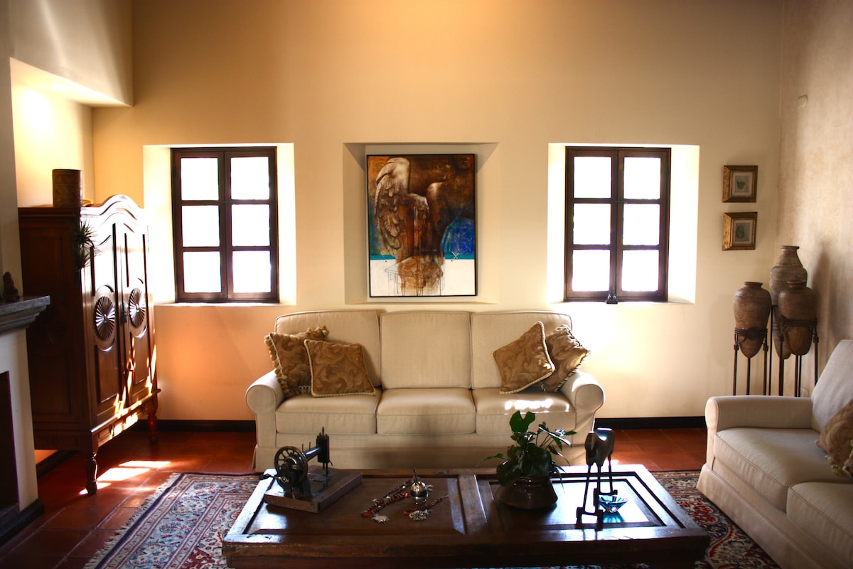 The living area features a light-colored sofa and armchairs, creating a comfortable seating arrangement. Two windows allow natural light to flood the space, revealing a vibrant wall art piece. An intricate wooden table lies in the center, accompanied by decorative accessories and plants.