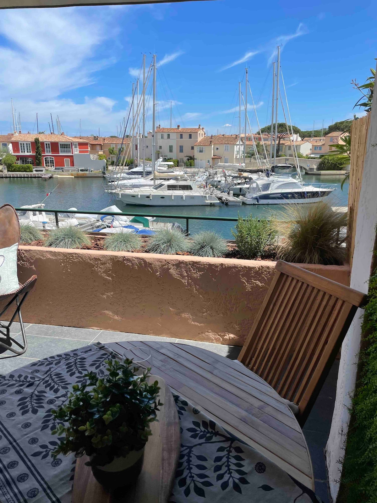 A terrace view features a wooden table and chairs, with a lush plant centerpiece. The backdrop showcases a marina filled with sailboats and yachts, framed by colorful buildings along the waterfront, under a clear blue sky.