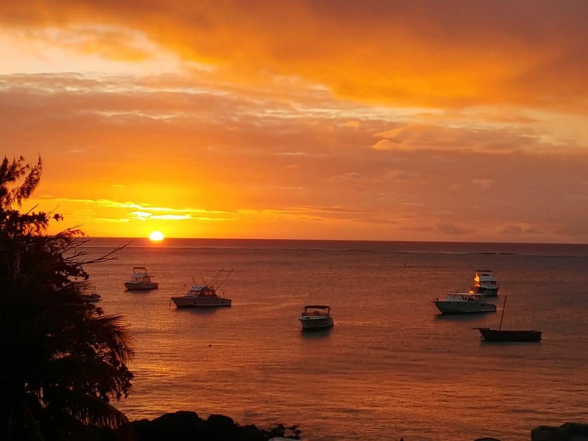 The image captures a vibrant sunset over the ocean, with several boats gently floating on the water. The sky is filled with warm hues of orange and yellow, reflecting on the sea's surface, while silhouettes of trees on the edge create a serene foreground.