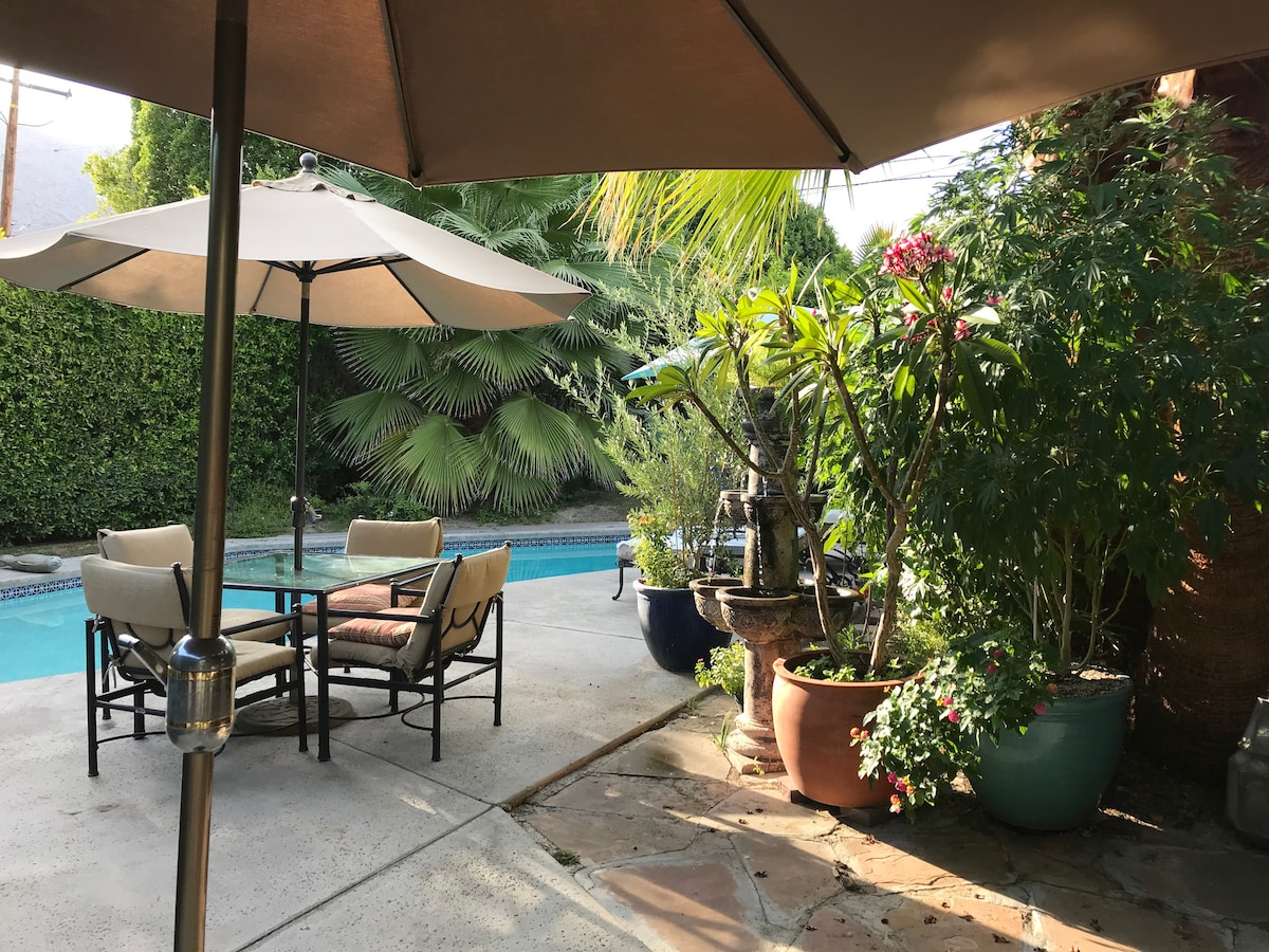 A serene poolside area is framed by lush tropical plants and vibrant flowers. Two lounge chairs with an umbrella provide shaded seating, while a water fountain adds a soothing element. The smooth patio is surrounded by greenery, enhancing the tranquil outdoor space.