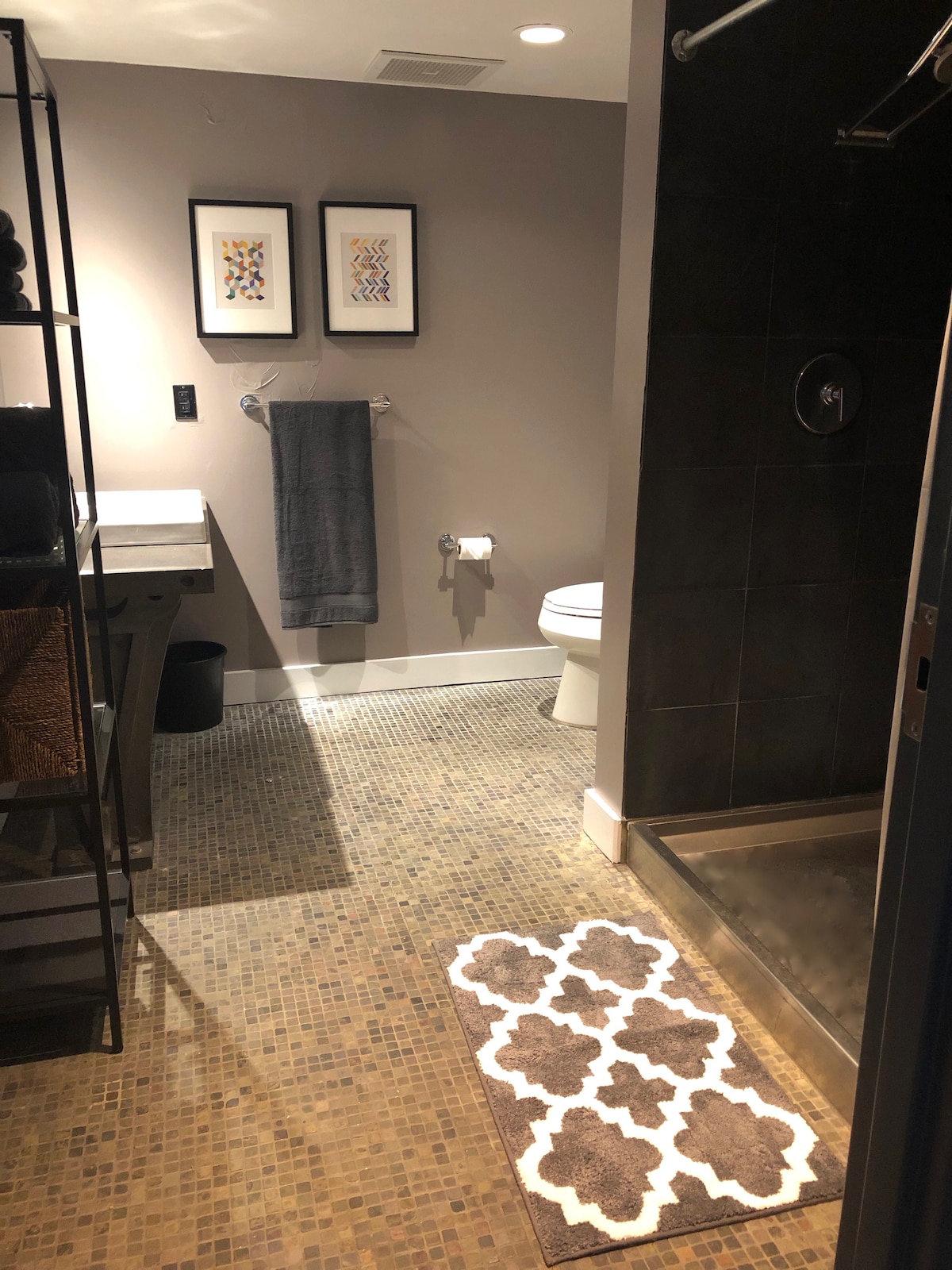 The bathroom features a modern design with a large, walk-in shower and a mosaic tiled floor. A dark towel hangs on the wall next to a framed artwork. A small area rug and shelves with neatly arranged towels and toiletries complete the space.