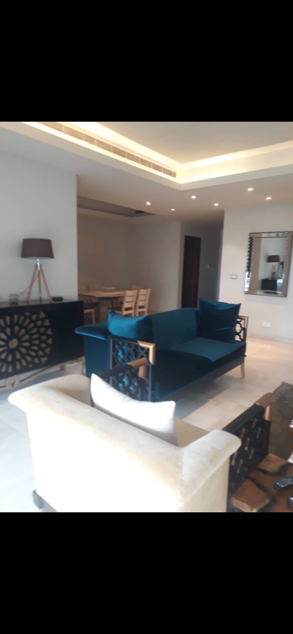 A spacious living area features a blue sofa with plush cushions and an elegant accent chair. Neutral-toned chairs and a dining space are visible in the background, complemented by soft lighting and mirrored decor, creating an open and inviting environment.
