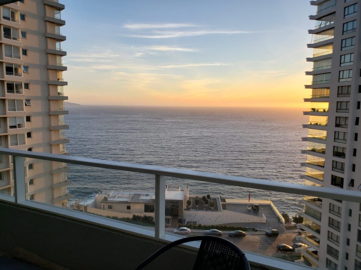 The expansive terrace features a clear view of the ocean, with the sun setting on the horizon. Surrounding high-rise buildings frame the scene, and the gentle waves can be seen in the distance, creating a serene coastal atmosphere.