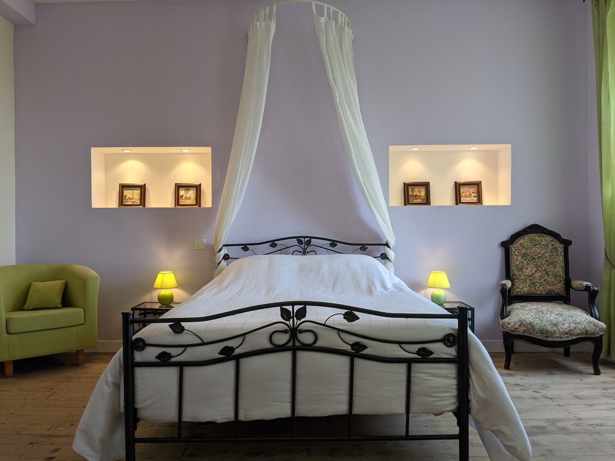 A spacious bedroom features a metal bed with a canopy and white bedding. Soft green lamps illuminate the nightstands. Two decorative wall niches display framed images. A green armchair and an elegant patterned chair add character, with light walls creating a calm environment.