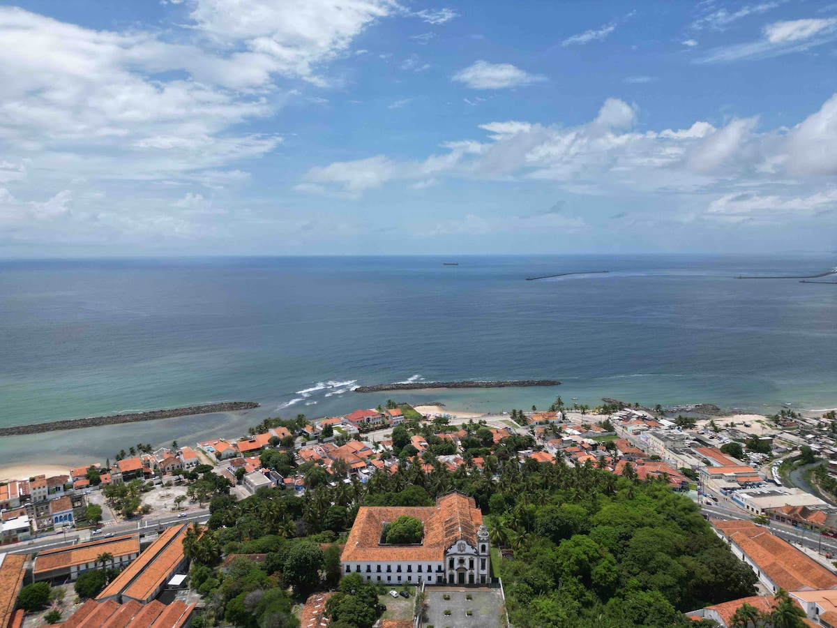 A panoramic view showcases the coastline meeting the ocean, dotted with small boats in the distance. Lush greenery blends with the terracotta rooftops of the town below, while the horizon stretches under a partly cloudy sky, offering glimpses of urban development along the shoreline.