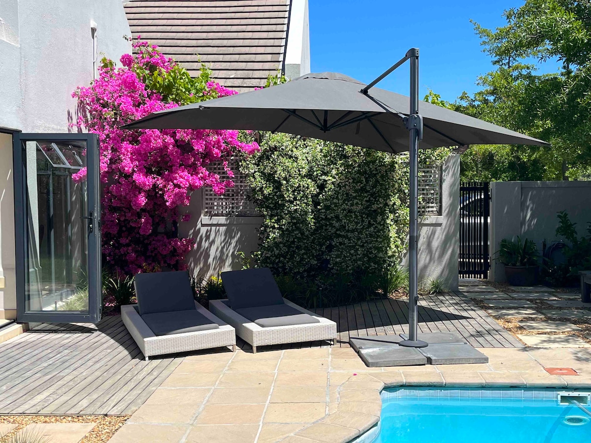 A serene outdoor area is featured, showcasing two sun loungers beneath a large umbrella, positioned beside a sparkling pool. Vibrant bougainvillea flowers in shades of pink provide a colorful backdrop, while lush greenery adds a touch of privacy to the space.