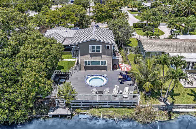 An aerial view captures a stylish home surrounded by lush greenery, featuring a large deck with reclining lounge chairs and a small pool. The property offers a serene lakeside setting, with tropical plants and an umbrella providing shaded areas for relaxation.
