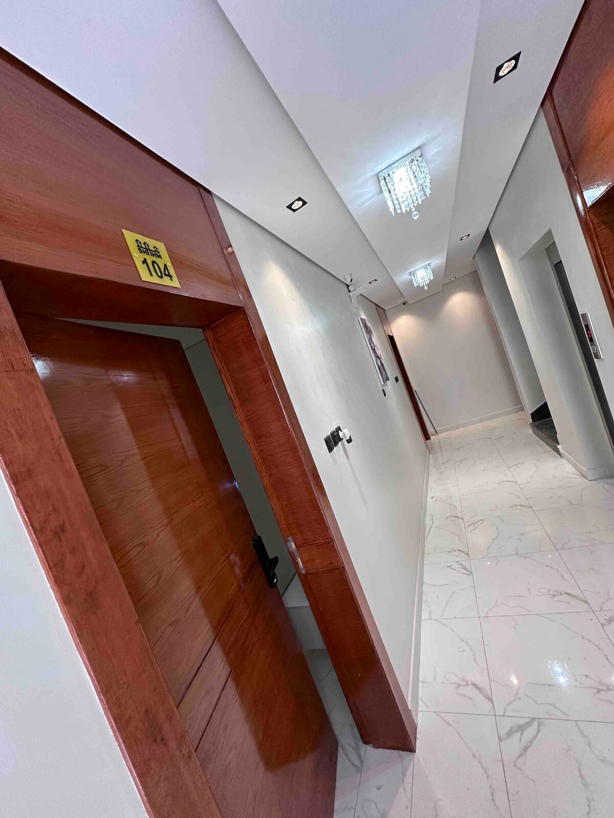 A well-lit hallway is lined with smooth, polished tiles. Wooden doors, marked with room numbers, are positioned along the walls. Subtle lighting fixtures and a crystal chandelier add a touch of elegance to the space.