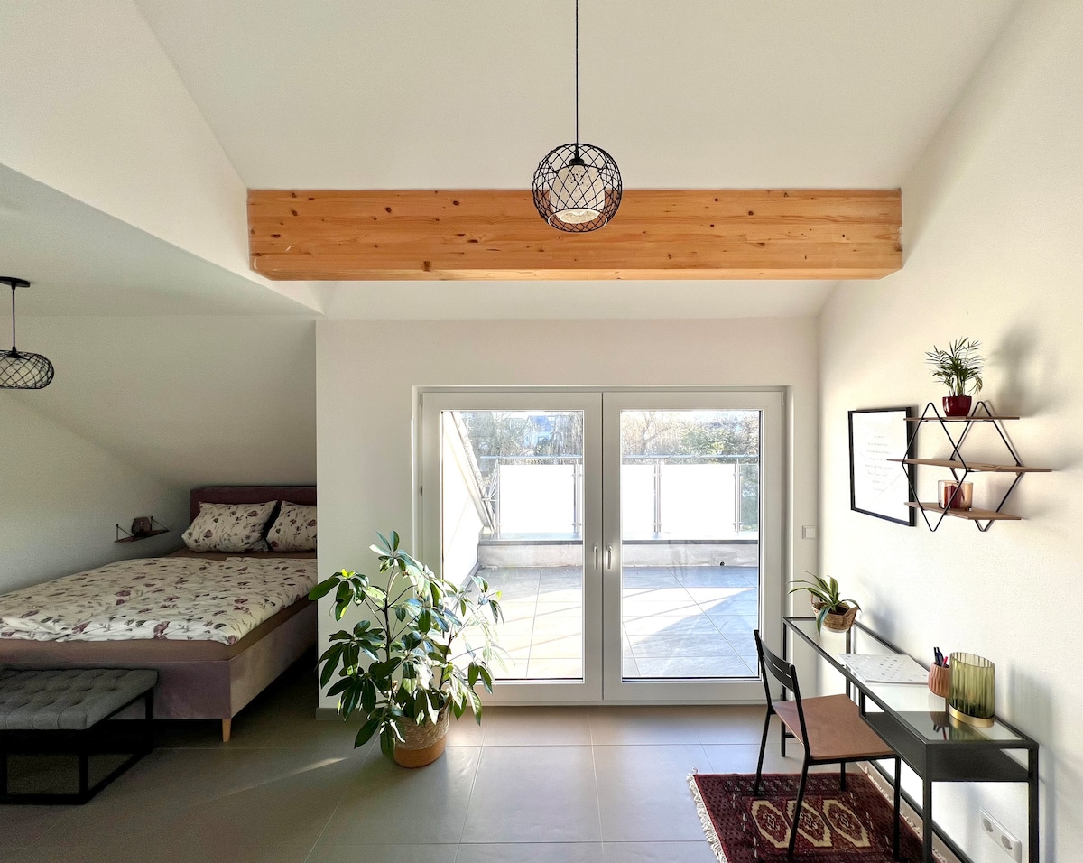 A cozy sleeping area features a spacious bed with a floral-patterned duvet. Natural light pours in through large glass doors that open to a private terrace. A small desk and chair, complemented by a decorative plant, offer a functional workspace.