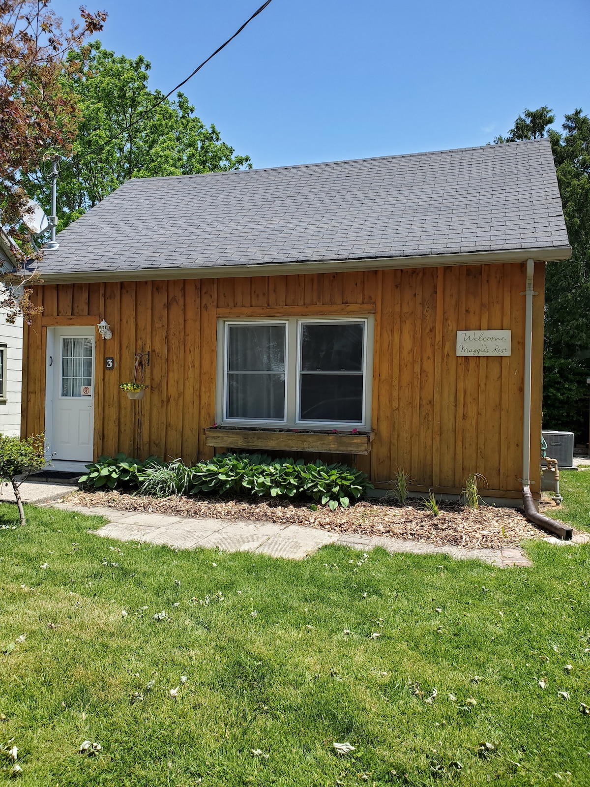 A charming cottage exterior features warm wooden siding and a sloped roof. Two large windows invite natural light, framed by lush greenery and flower beds. A small sign is mounted next to the door, identifying the property, while a pathway of stones leads to the entrance.