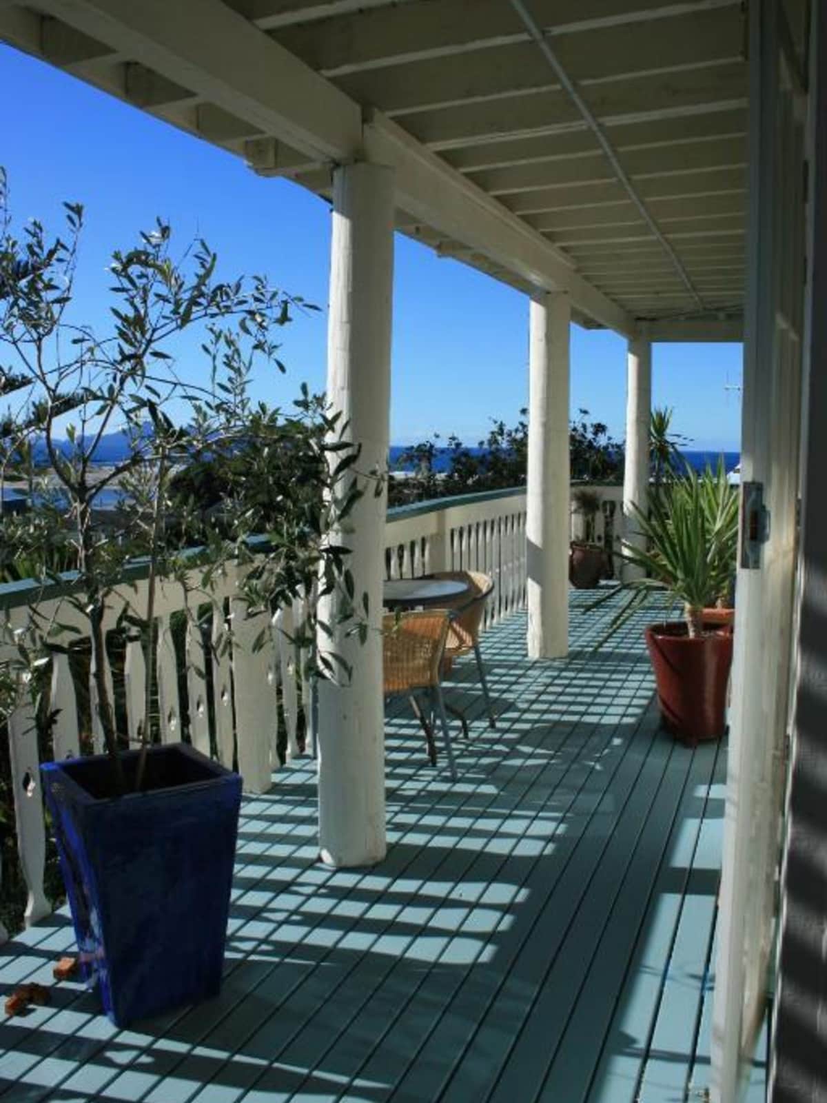 A private veranda is highlighted by sea views, featuring potted plants and outdoor furniture. Sunlight casts distinct shadows on the blue-painted deck, complemented by the white railings and columns that frame the space.