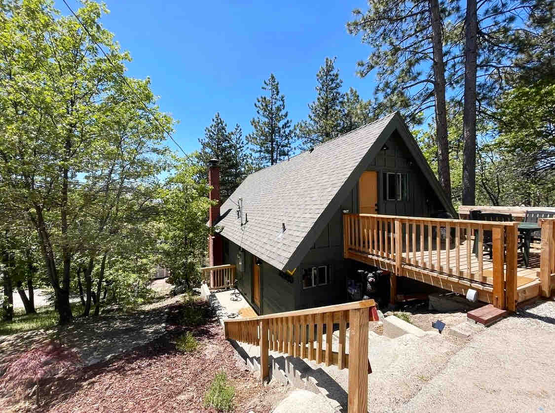 The cabin features a sloped roof and wooden patio, surrounded by tall pine trees. A staircase leads to the entrance, with a pathway that connects to the driveway. Bright sunlight illuminates the exterior, highlighting the natural setting.