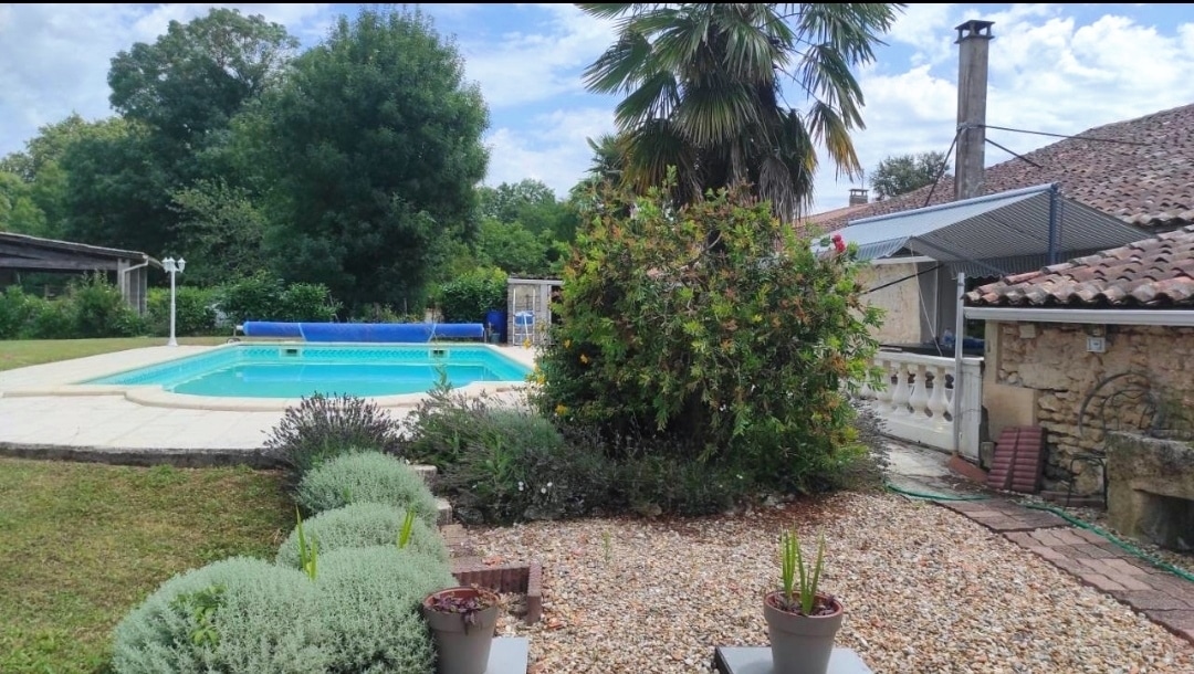 A private outdoor area features a swimming pool surrounded by well-maintained grass. Lush trees and palms provide shade while colorful flowers and low shrubs enhance the garden. A stone structure is visible nearby, with an outdoor dining area and lounge chairs arranged for relaxation.