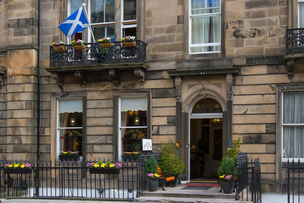 The entrance of the Edinburgh Thistle Hotel features traditional stone architecture with large windows adorned with flower boxes. A Scottish flag is displayed prominently, while potted plants enhance the welcoming atmosphere. A decorative wrought-iron railing surrounds the patio area.