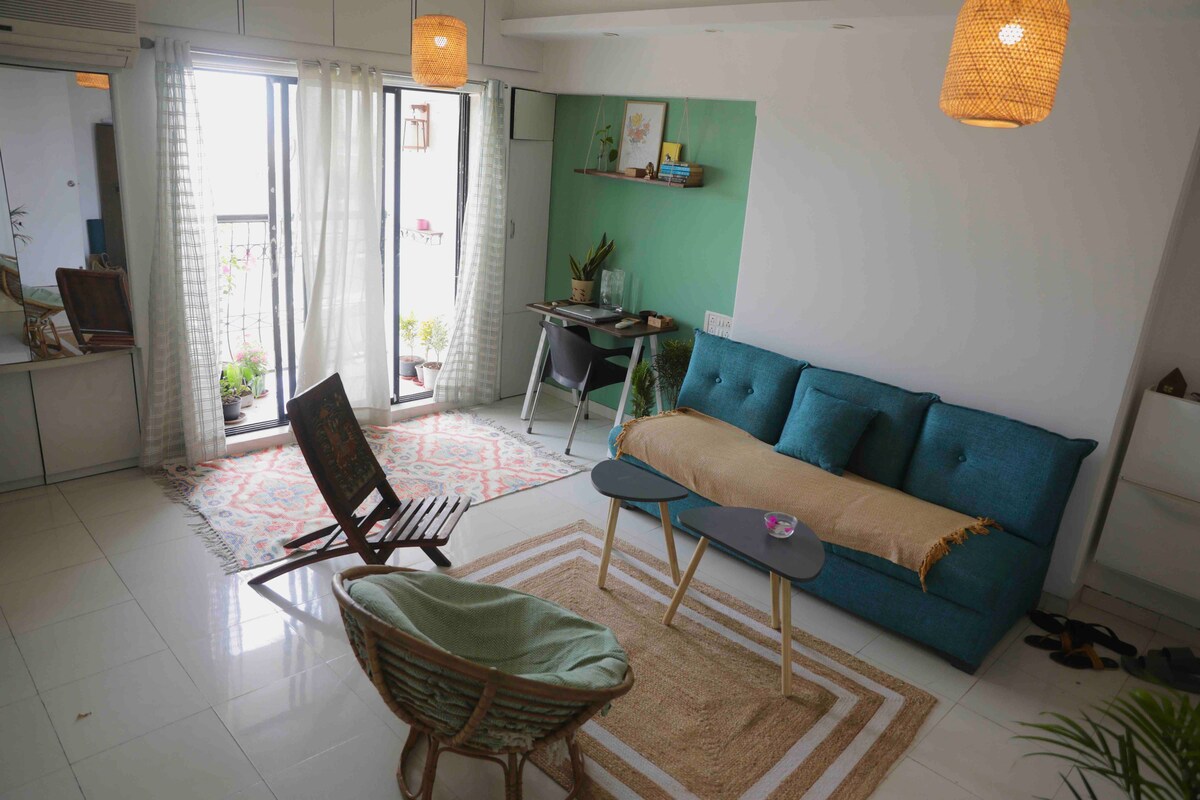 A bright living area is showcased with large glass doors leading to a small balcony adorned with plants. A comfortable teal sofa is situated across from a woven chair and a low table. Natural light floods the space, enhancing the sleek tiles and decorative rug.