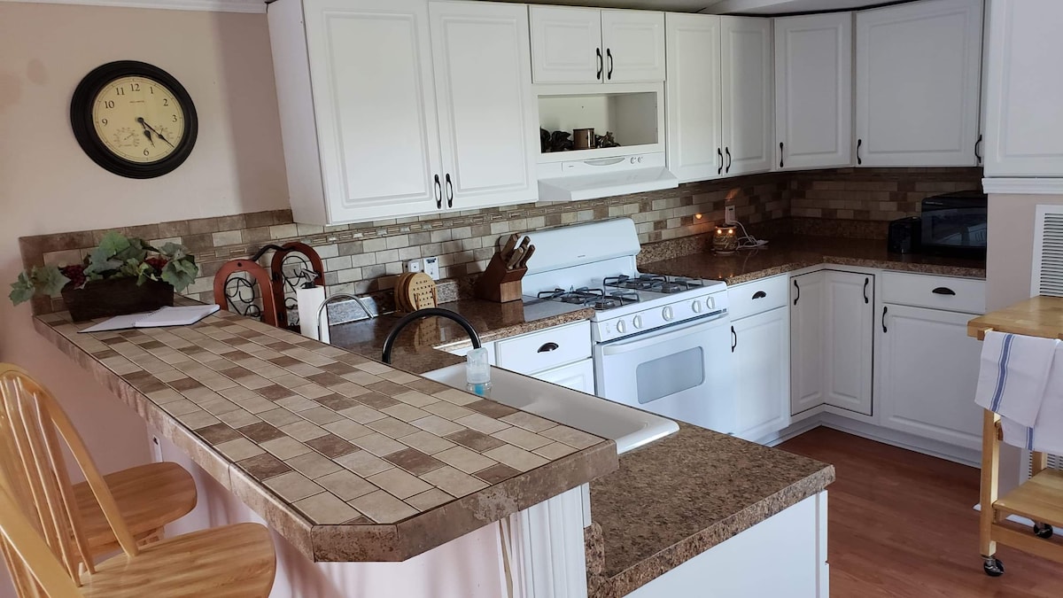 A modern kitchen features white cabinetry and a spacious layout. A tiled breakfast bar is highlighted, with wooden stools positioned nearby. Essential appliances include a gas stove and a microwave, while decorative items and a clock add warmth to the space.