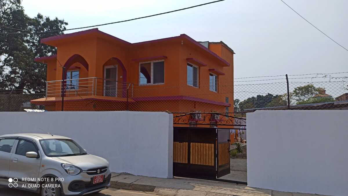 The exterior of a vibrant orange building is visible, featuring a modern design with arched balconies and large windows. A gated entrance is present at the front, alongside a white wall that provides privacy. Lush greenery surrounds the property, enhancing the inviting atmosphere.