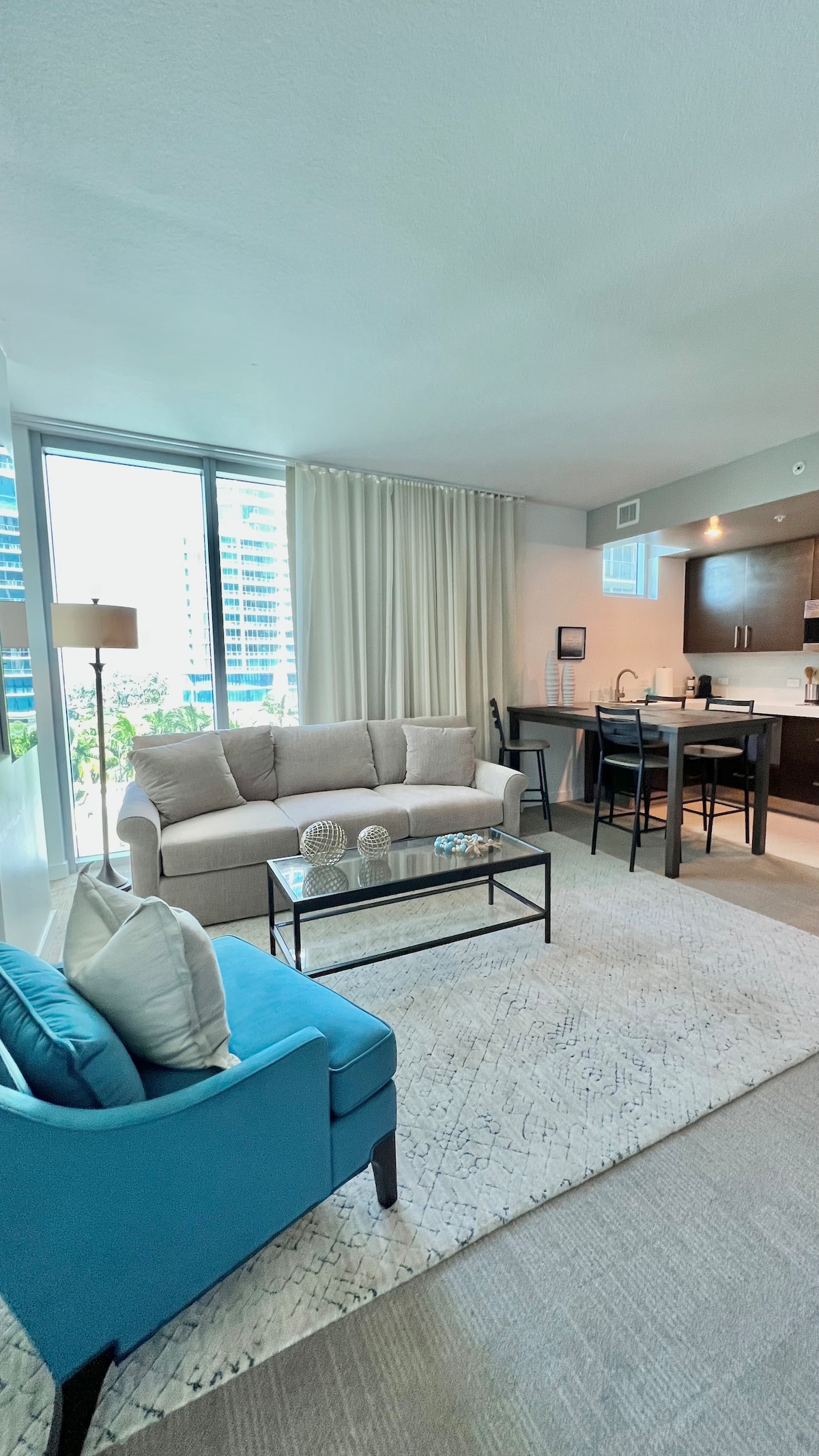 A modern living space features a light-colored sofa accompanied by a blue accent chair. A glass coffee table sits in the center, displaying decorative items. The kitchen, visible in the background, includes dark cabinetry and a dining area with four chairs. Large windows allow natural light to brighten the room.