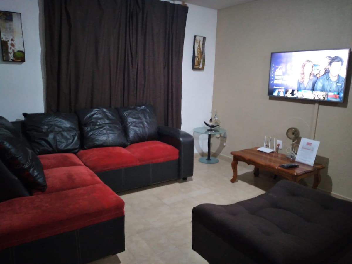 A comfortable living room is showcased, featuring a dark sofa with red accents and a matching lounge chair. A television is mounted on the wall, displaying content. Soft lighting illuminates the space, complemented by decorative wall art and sheer curtains.