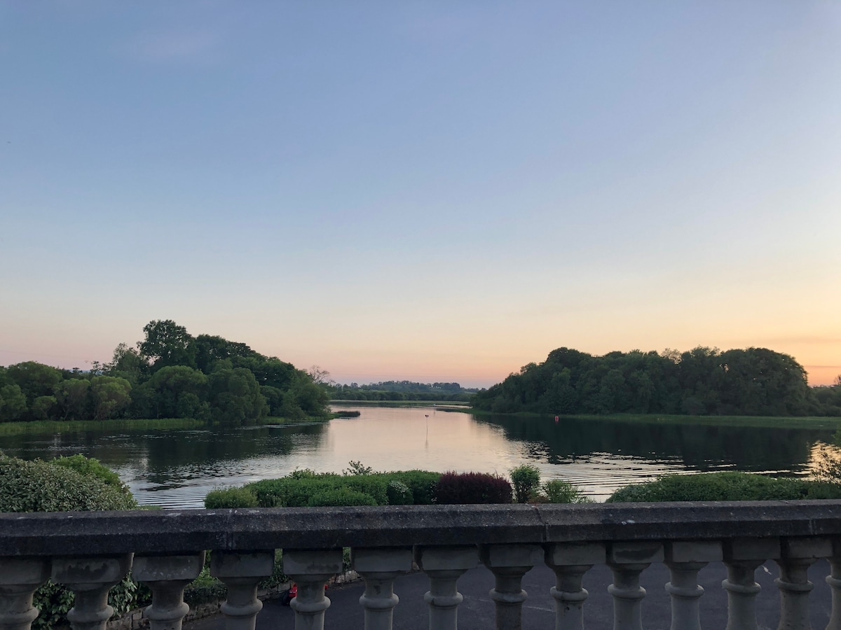 A serene view of the lake is captured from a stone balcony. Lush greenery surrounds the water, reflecting the soft hues of the evening sky. The horizon is gently illuminated, suggesting a peaceful transition from day to night.