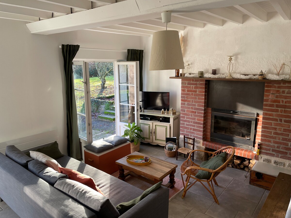 A cozy living area is depicted, featuring a comfortable sofa and a wooden coffee table. A brick fireplace adds warmth, while large windows allow natural light to fill the space. A television is positioned across from the seating, and an inviting outdoor view is glimpsed through the open doors.