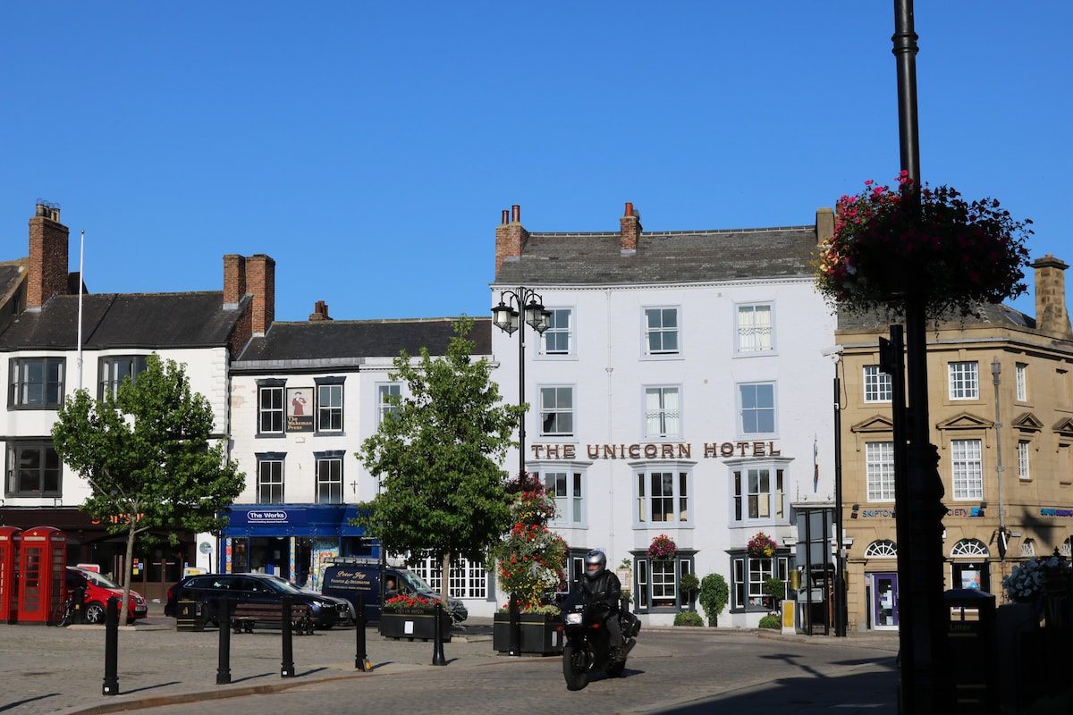 A vibrant streetscape showcases the historic buildings of Ripon, including the prominently displayed Unicorn Hotel. Trees and hanging flower baskets line the square, with parked cars and a motorcycle noticeable on the street. Clear blue skies complement the scene, enhancing the lively atmosphere.