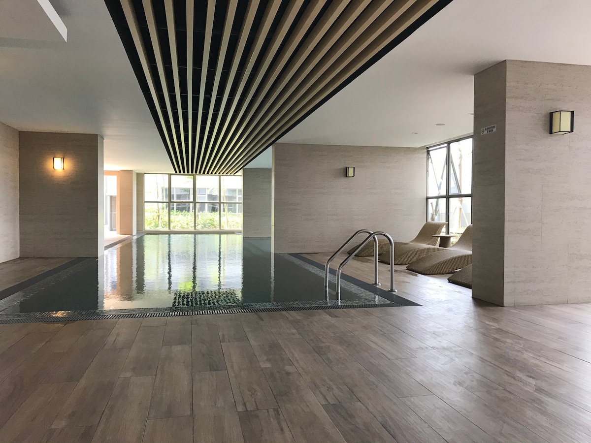 The indoor swimming pool area features a sleek design with wooden slats adorning the ceiling. Lounge chairs are situated near the pool, complemented by large windows that allow natural light. The serene water surface reflects the surrounding contemporary architecture.