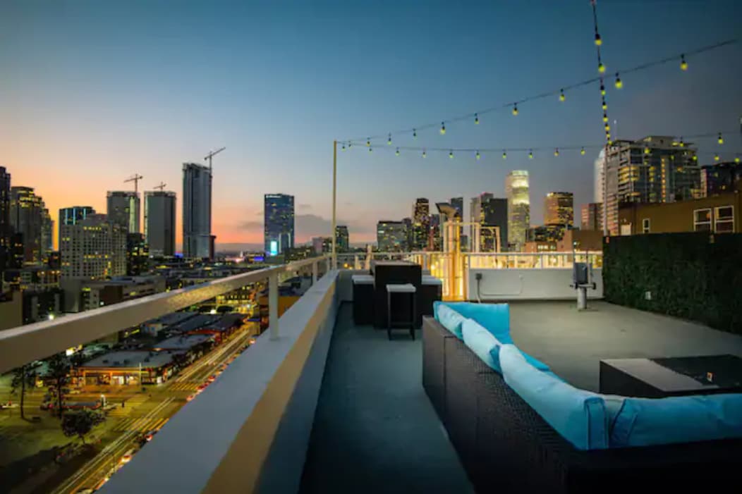 A private rooftop terrace is showcased, featuring modern outdoor furniture and string lights that offer a warm glow against the backdrop of the city skyline. The setting sun casts soft hues across the skyline, enhancing the urban view.