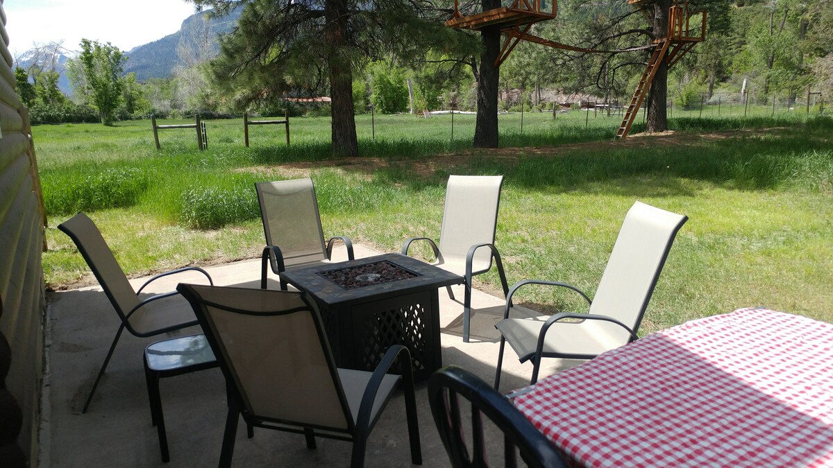 An outdoor seating area is featured on a concrete patio, surrounded by lush green grass. Four light-colored chairs are positioned around a gas fire table. A red and white checkered tablecloth covers a nearby table, providing a space for outdoor dining or relaxation.