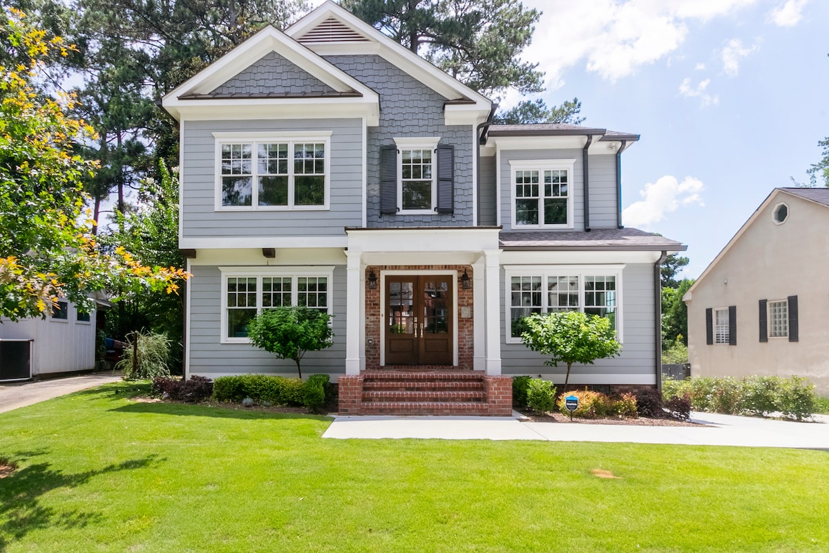 The exterior of a two-story home is shown, featuring a light gray facade with dark green shutters. The entrance is marked by a welcoming porch with a set of stairs leading to double doors. Fresh greenery and landscaped shrubs surround the house, contributing to a pleasant outdoor setting.