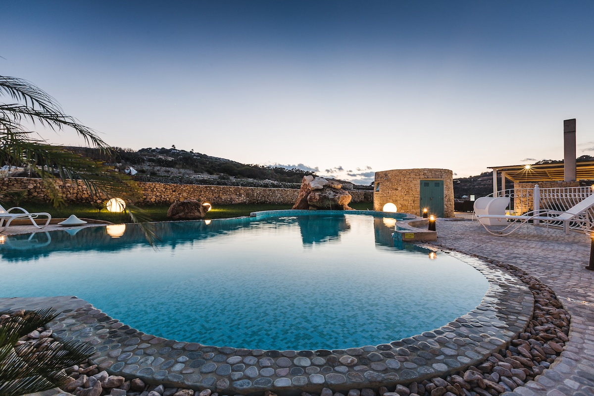 A tranquil outdoor area is depicted with a spacious swimming pool reflecting the evening sky. Surrounding the pool are smooth stones and comfortable lounge chairs. A natural rock formation adds a touch of charm, while soft outdoor lighting creates a calming ambiance.