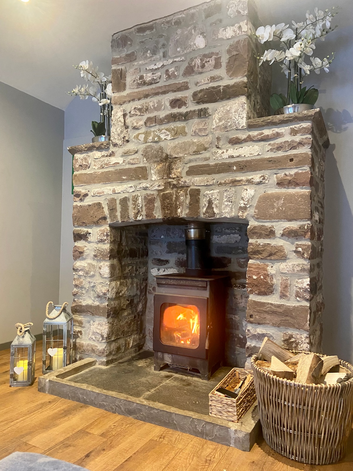 A rustic stone fireplace serves as the focal point, featuring a cast iron wood burner with a visible flame. Two lanterns are placed on the left, while a woven basket filled with logs sits to the right. Decorative white orchids are positioned atop the mantel.