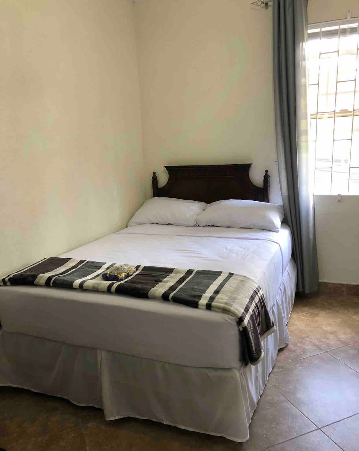 A sleeping area features a neatly made bed with a light-colored quilt and a decorative throw blanket. Natural light enters through a window adorned with sheer curtains, illuminating the room's neutral walls and tiled floor.