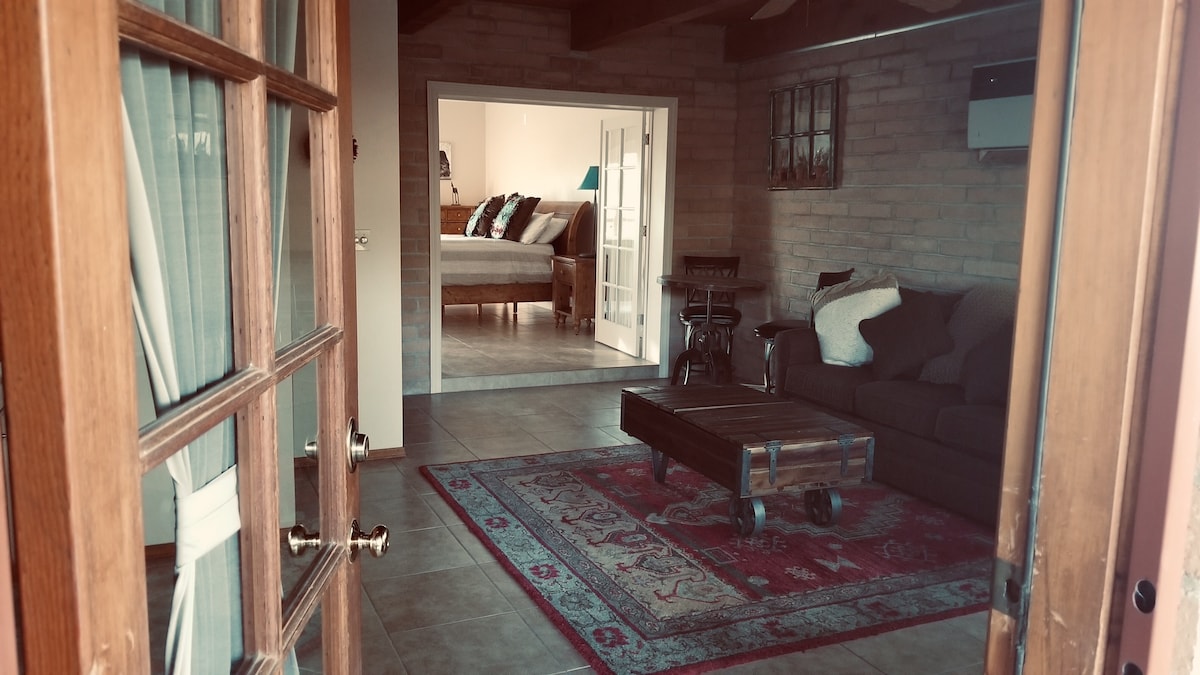 A welcoming entrance reveals a cozy living room showcasing a spacious sofa, a rustic coffee table, and warm tones. Beyond, a second sleeping area is partially visible, complemented by light-colored furnishings and a comfortable atmosphere.