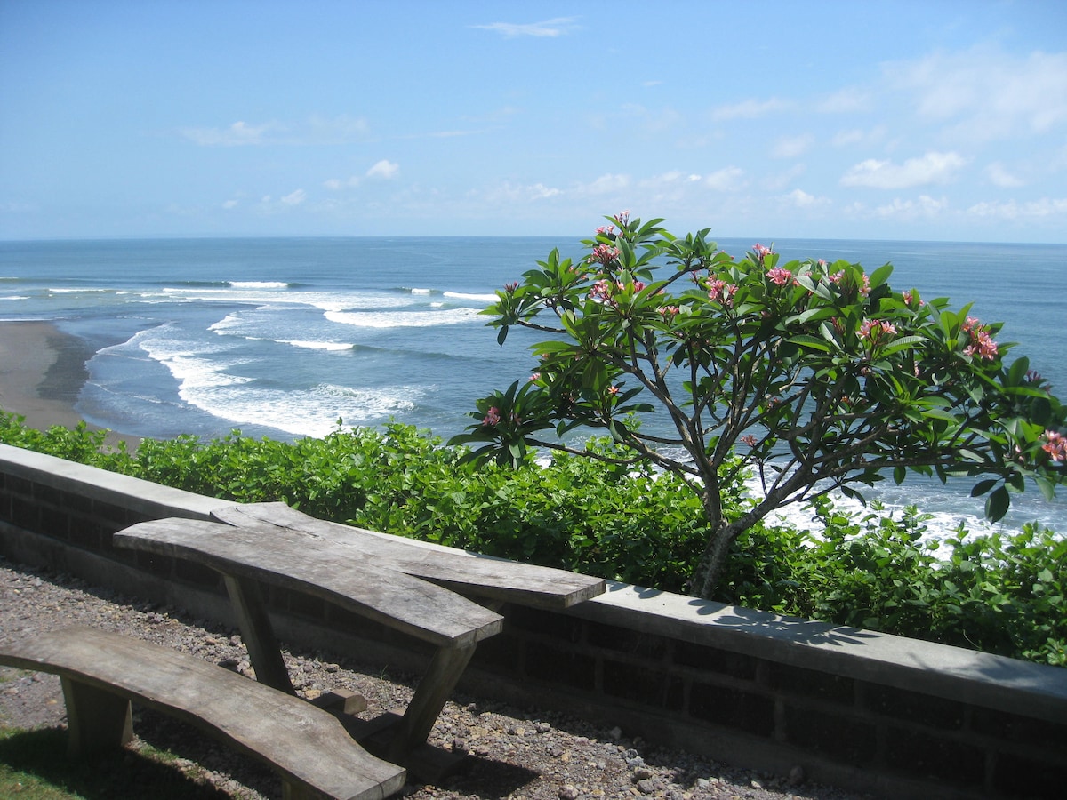 A wooden picnic table is positioned near a low stone wall, overlooking a serene beach. Gentle waves roll onto a secluded black sand shoreline, framed by lush green foliage and a flowering tree. A clear blue sky stretches above, complementing the tranquil ocean view.