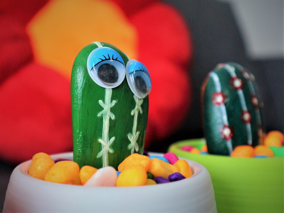 Two painted rock cacti with googly eyes are displayed in white and green pots filled with colorful pebbles. A plush red flower is visible in the background, adding a playful touch to the setting.