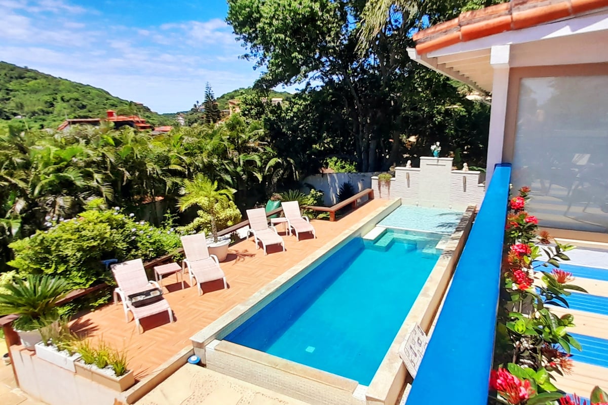 A modern pool area is surrounded by lush greenery, with sun loungers positioned along the tiled terrace. The pool features a sleek design, with a shallow area and clear water inviting relaxation under the sunny sky.