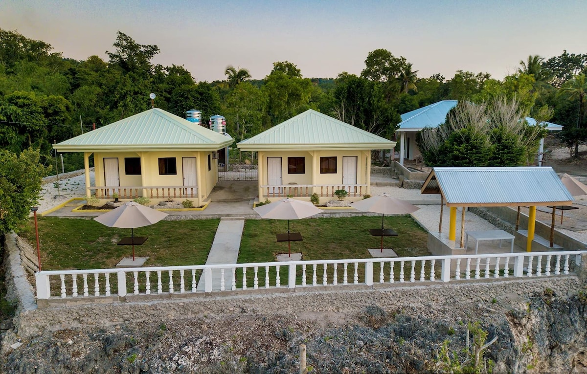 Two well-constructed cottages with light yellow exteriors are shown, set in a lush, green landscape. Outdoor seating areas with umbrellas are visible in front of each unit, providing shaded spots for relaxation. A pathway connects the cottages, leading toward a nearby pavilion.