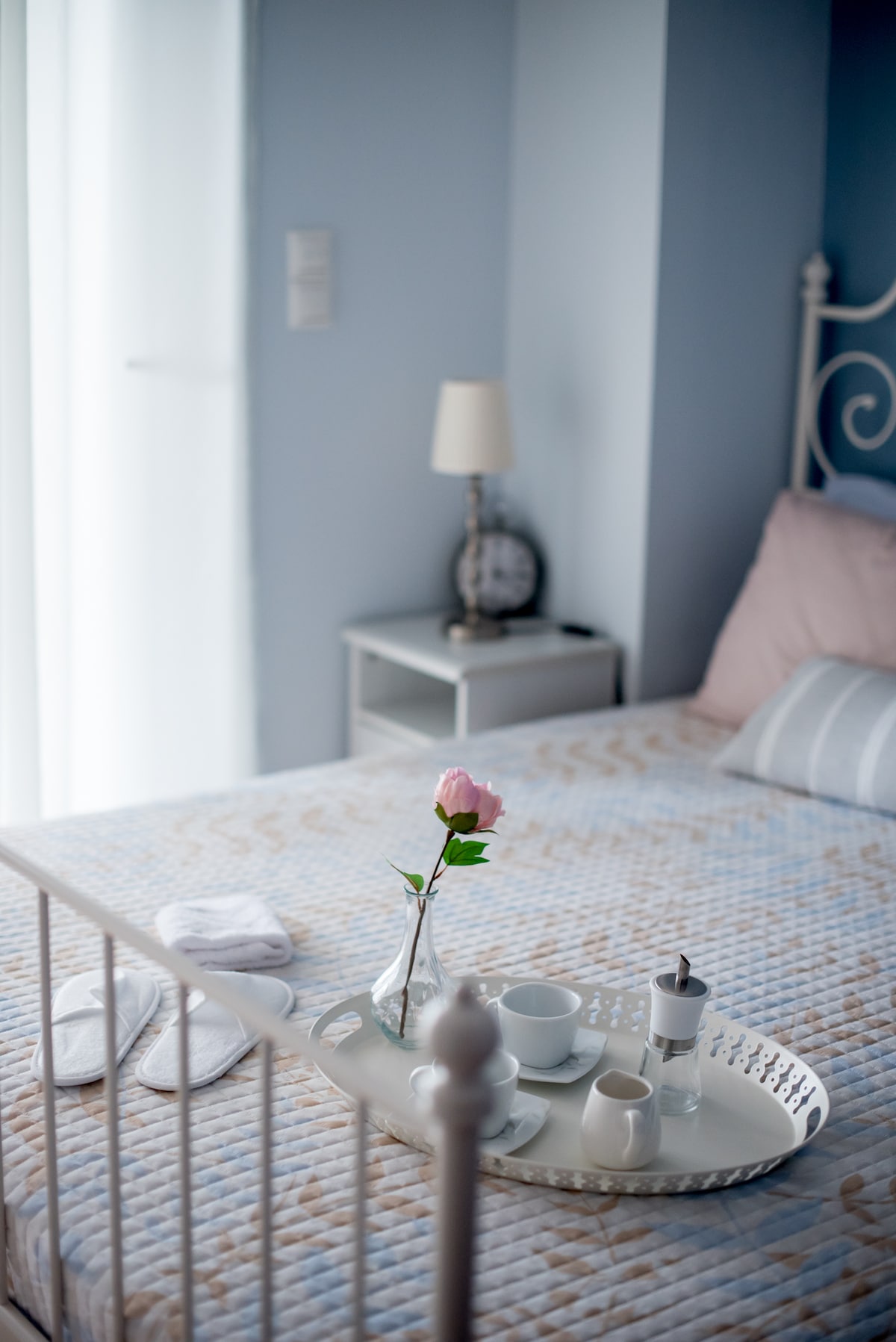 A light-filled bedroom features a spacious bed adorned with a quilted cover. A bedside table holds a clock and a lamp. A tray with a flower, cups, and a small carafe is positioned on the bed, enhancing the tranquil setting.