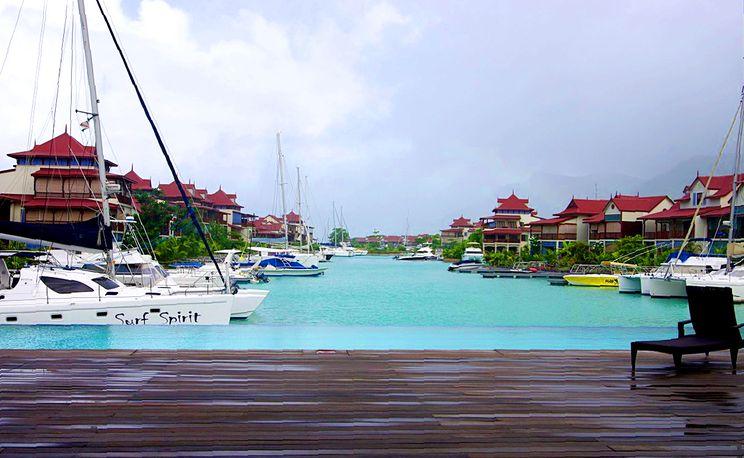 The serene marina features several yachts docked along the water, with a backdrop of lush green hills and buildings displaying traditional architecture. Calm turquoise waters reflect the overcast sky, providing a tranquil atmosphere. A wooden deck is visible in the foreground, enhancing the scenic view.
