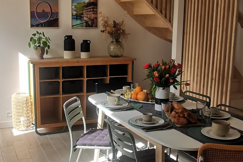 A dining area is furnished with a large table set for several. Fresh flowers, fruit, and pastries are placed on the table. Two large vases are centered on a sideboard, while natural light filters in, highlighting the wooden staircase in the background.
