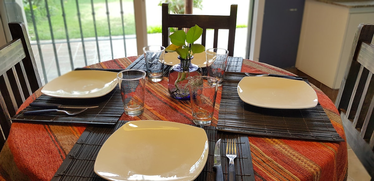 A dining table is set with four white plates, accompanied by glasses and utensils. A decorative centerpiece with green leaves is placed in a blue vase. The tablecloth features an orange and red pattern, complementing the natural light coming from a nearby window.