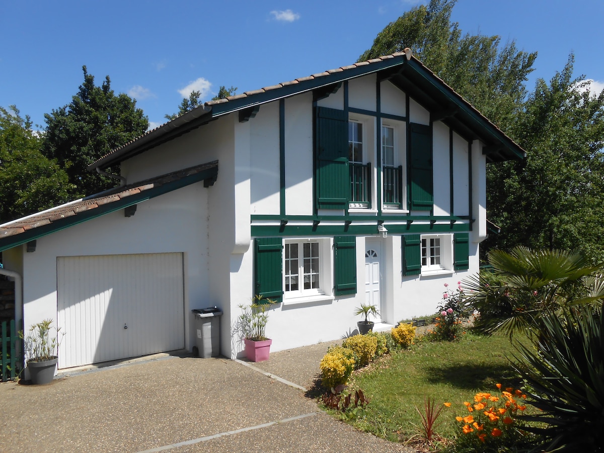 The exterior of the house presents a two-story design with green shutters and white walls, complemented by a driveway lined with flowering plants. A small garage is visible on the left side, surrounded by lush greenery.