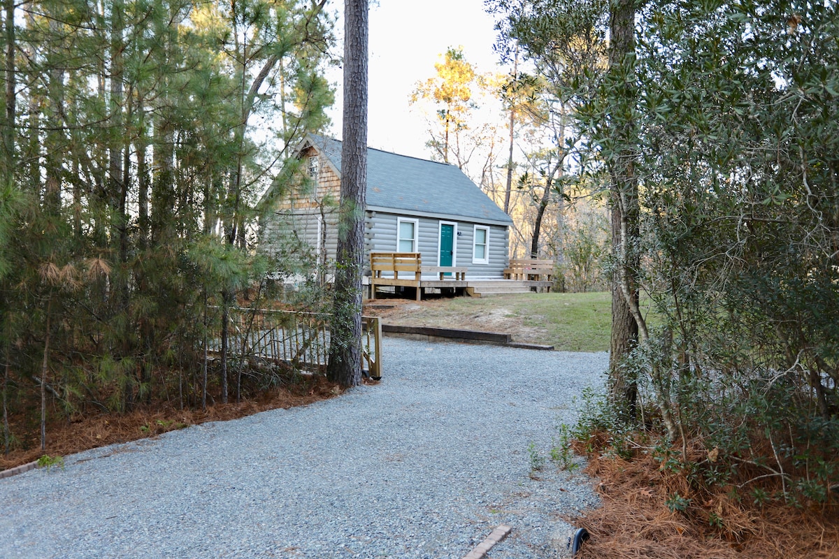 A cabin is nestled among trees, featuring a gravel driveway leading to a spacious yard. The structure has light-colored siding and a blue door, with a wide front porch accessible from the driveway.