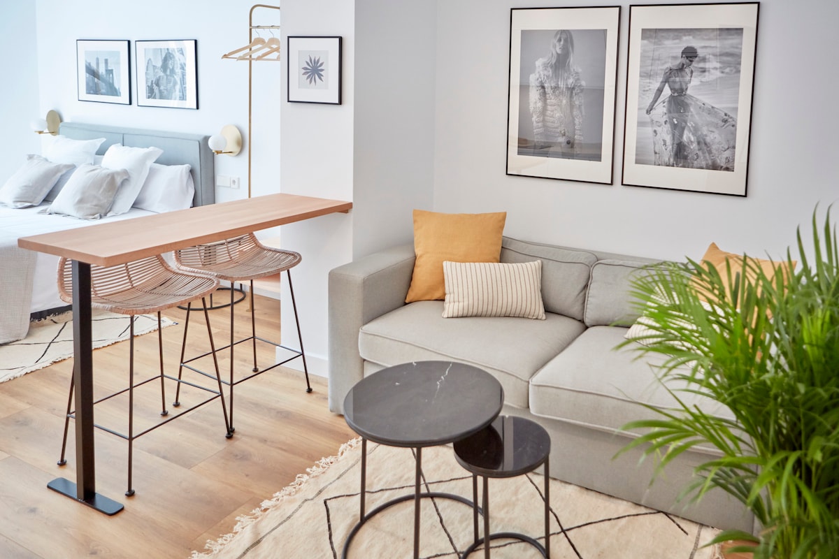 A serene living area features a light gray sofa with a yellow accent pillow, complemented by two round coffee tables. A sleek dining table with three bar stools is positioned nearby, and black-and-white artwork adorns the walls, enhancing the modern aesthetic.
