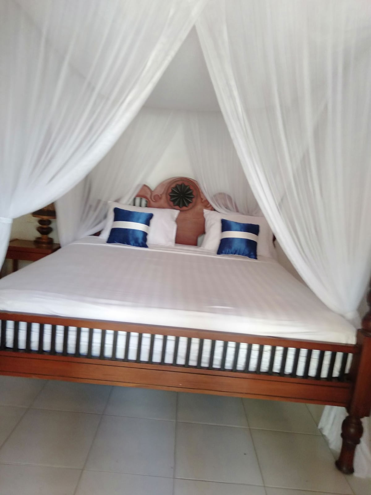 A spacious bed is draped with a white canopy, providing a serene sleeping environment. Two decorative pillows with blue accents are placed against the headboard. The room features neutral tile flooring and wooden side tables, contributing to a calm and restful atmosphere.