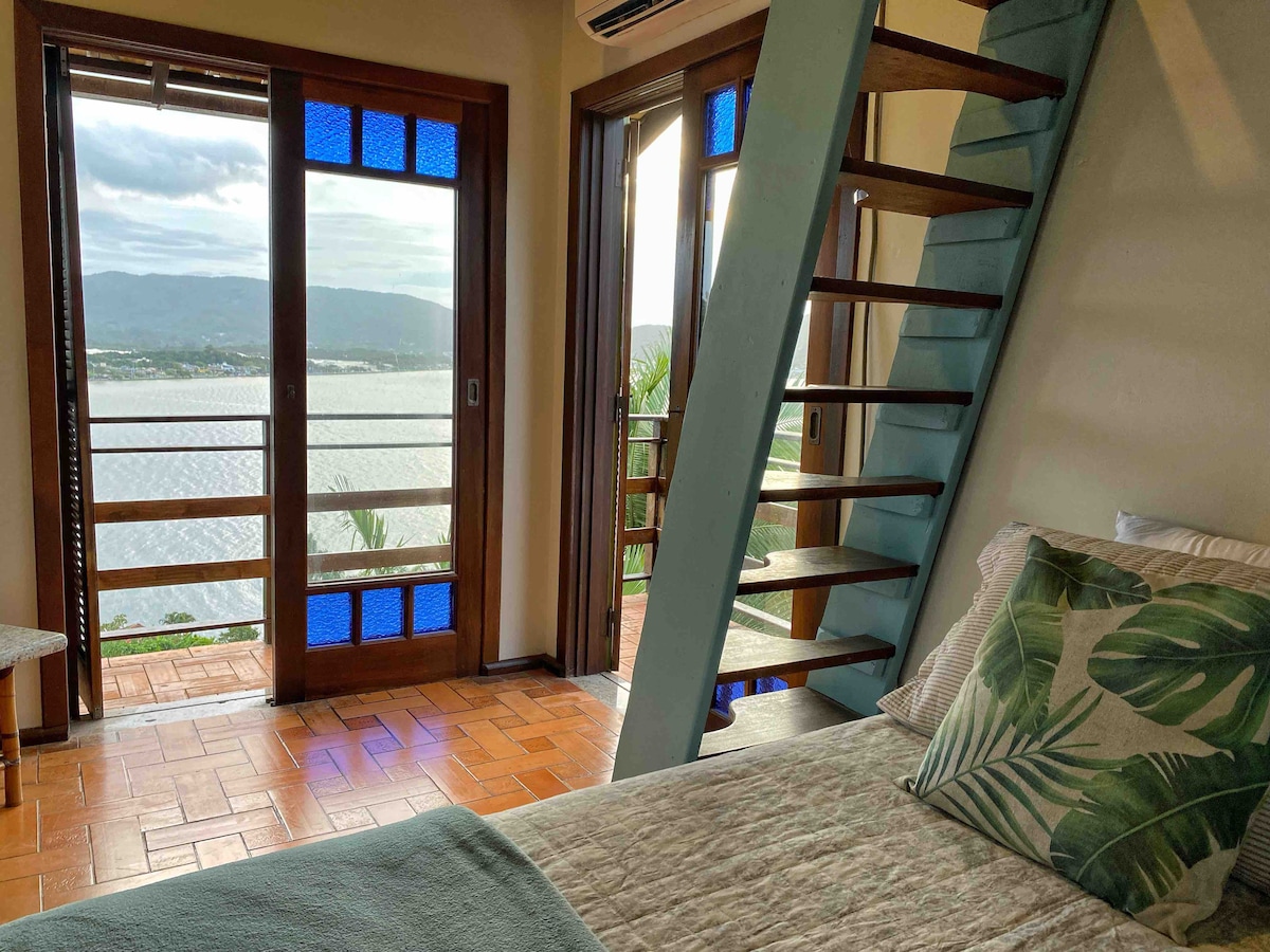 A sleeping area features a spacious bed with a light, neutral cover and decorative pillows. Large glass doors provide a view of the lake and mountains, while a wooden staircase leads to an upper level. Natural light fills the room, enhancing its warm ambiance.
