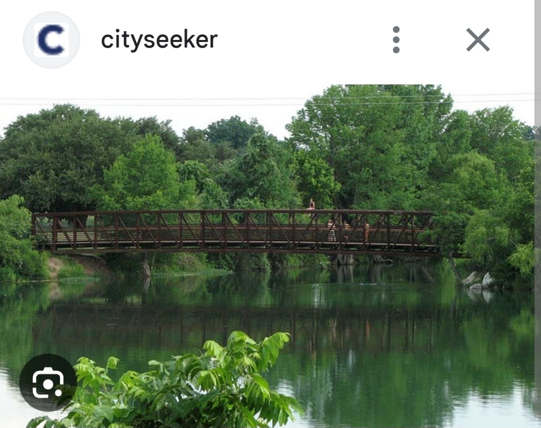 A sturdy metal pedestrian bridge spans a tranquil body of water, surrounded by lush green trees and foliage. The reflections of the trees and bridge can be seen in the calm water, creating a serene natural setting.