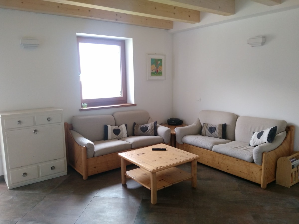 A spacious living area features two light-colored sofas with decorative cushions and a wooden coffee table centered between them. A bright window allows natural light to fill the room, while a small sideboard adds to the functional aesthetics.
